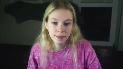 Media: Video of a blonde Caucasian girl with straight hair, wearing a pink floral shirt, sitting indoors. Background features a dark sofa and a partially visible green blanket.