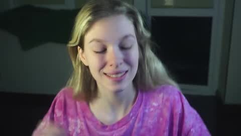 Media: A video of a young girl with fair skin and long blonde hair, wearing a pink tie-dye shirt, smiling with eyes closed. The background features a dark room with a green cloth hanging.