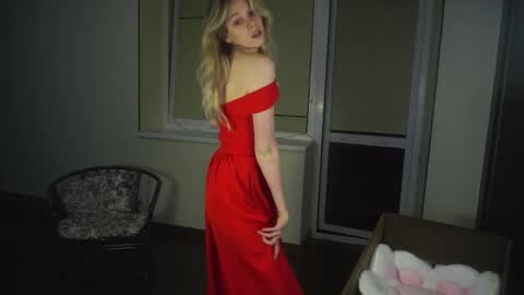 Media: Video of a blonde woman in a red off-shoulder dress, standing in a dimly-lit room with a glass door, a patterned chair, and a cardboard box.