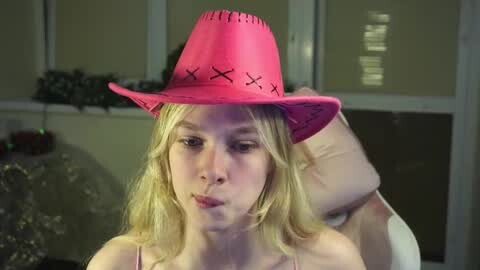 Media: Video of a blonde woman with fair skin wearing a bright pink cowboy hat with black cross stitching, blowing a bubble in a dimly lit room with closed white blinds and a decorative Christmas wreath.