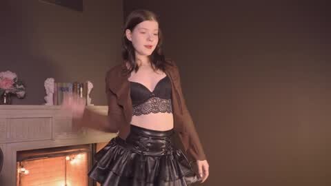 Media: Video of a young woman with light skin and straight dark hair, wearing a black lace bra and brown jacket over a black leather skirt, standing in a dimly lit room with a fireplace and bookshelves.