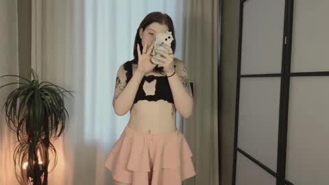 Media: Video of a slim, pale-skinned young woman with shoulder-length dark hair, wearing a black lace bra and a short, frilled pink skirt, taking a mirror selfie. Background includes a potted plant, a shoji screen, and curtains.