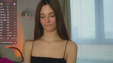 Media: A video of a young woman with long brown hair, fair skin, and a thin build, wearing a black spaghetti strap top, standing in a softly lit bedroom.