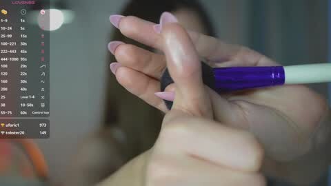 Media: Video of a woman with fair skin, long dark hair, manicured nails painted lavender, holding a purple makeup brush, blurred background, digital overlay showing makeup tutorial.