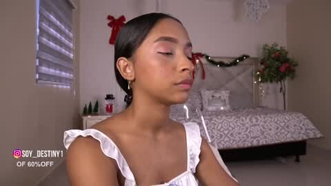 Media: Video of a serene, dark-skinned woman with long black hair in a bun, wearing a white ruffled blouse, in a cozy bedroom with a bed, festive decor, and a window.
