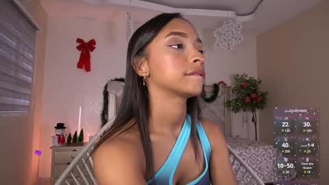 Media: Video of a young, dark-skinned woman with long black hair, wearing a blue one-shoulder dress, in a cozy bedroom with Christmas decorations, digital weather display on screen.