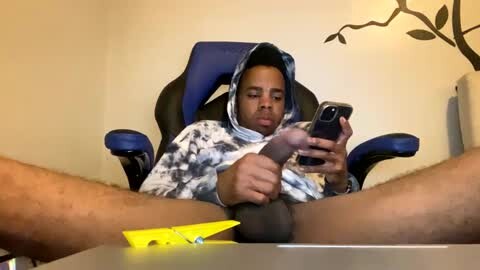 Media: Video of a young man with medium brown skin, wearing a tie-dye hoodie, sitting on a blue gaming chair. He's holding a smartphone and masturbating with a grey dildo. The room features a wall decal of tree branches.
