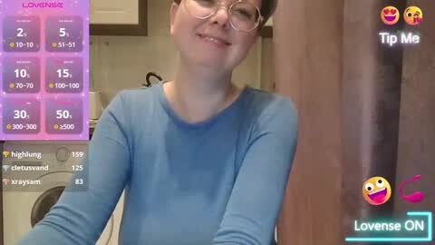 Media: Video of a young, fair-skinned woman with glasses and a blue shirt, smiling in a kitchen with a Lovense ON app overlay showing her stats.