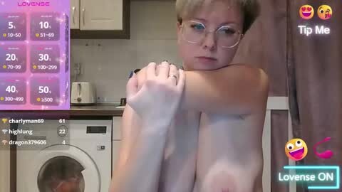 Media: A video of a fair-skinned, short-haired woman with glasses, sitting naked in a modern kitchen, arms folded, looking tired. Digital overlays show her stats and a \"Love Sense ON\" message.