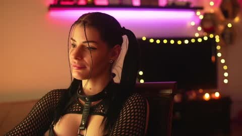 Media: Video of a woman with long dark hair in pigtails, wearing a black fishnet top and a leather harness, seated in a dimly lit room with string lights and a television in the background.