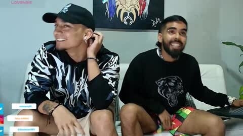 Media: Video of two smiling men, one in a black and white patterned shirt, the other in a black shirt with a tiger graphic. They're seated on a white couch, surrounded by vibrant, colorful cushions.
