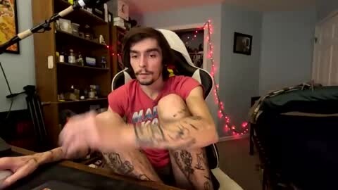 Media: Video of a young man with tattoos, sitting on a gaming chair in a dimly lit room with red fairy lights, wearing a red t-shirt and black shorts.