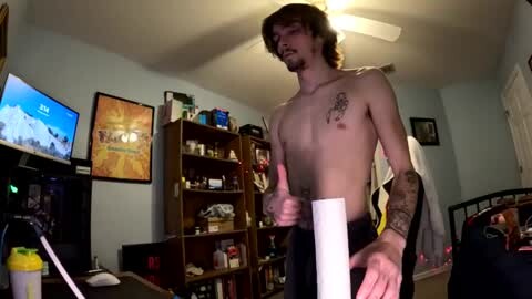Media: Video of a shirtless, slender young man with tattoos and medium-length brown hair giving a thumbs-up, standing in a cluttered, dimly lit living room with a TV, bookshelf, and posters on the walls.