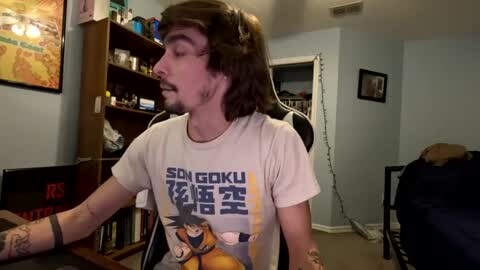 Media: Video of a man with shoulder-length brown hair, wearing a Dragon Ball Z T-shirt, sitting at a desk in a dimly lit room with bookshelves and a small dog.