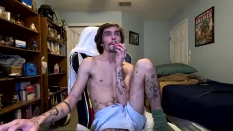 Media: Video of a shirtless, tattooed, slender young man with light skin, sitting on a gaming chair in a cluttered bedroom with shelves, posters, and a bed in the background.