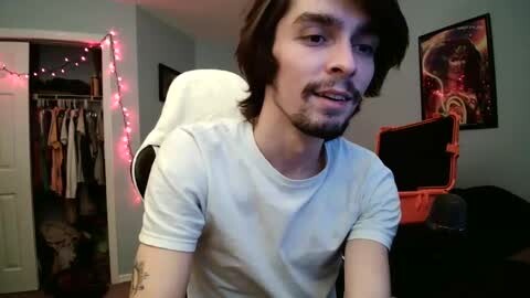 Media: Video of a young man with medium-length dark hair and a trimmed beard, wearing a white t-shirt, sitting in a room with a white chair, pink fairy lights, and a red guitar case.
