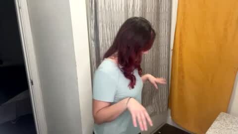 Media: Video of a woman with wavy, dark red hair, wearing a light blue t-shirt, standing in a bathroom with a silver shower curtain and mustard-yellow towel. She gestures with her hands.