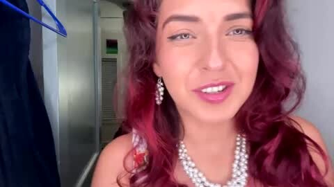 Media: Video of a smiling woman with long, wavy, deep red hair, wearing a pearl necklace and earrings, standing in a dimly lit hallway with a blue jacket hanging nearby.