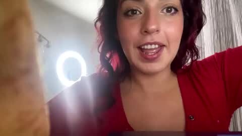 Media: Video of a smiling woman with wavy dark hair, wearing a red cardigan, in a modern bathroom with a circular mirror light.