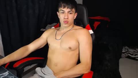 Media: Video of a young, shirtless, slim Latino man with short dark hair, sitting in a black and red gaming chair, wearing gray pants, with a black curtain backdrop.