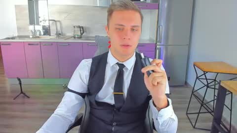 Media: Video of a young, light-skinned man with short, light brown hair, wearing a black suit, white shirt, and black tie, holding a microphone in a modern, brightly lit kitchen with pink and purple cabinets.