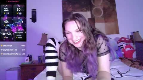 Video of a young woman with long, wavy hair, wearing a sheer black top, lying on a bed with a purple-striped blanket. The room features a wooden nightstand, a red plush toy, and a purple lamp.