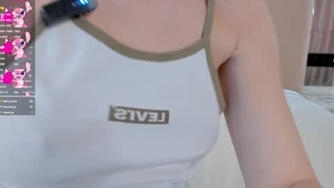 Media: A video of a fair-skinned woman wearing a white tank top with green trim and the word \"LEVIS\" on the chest. Her arm is raised, holding a smartphone with a pink screen displaying live stream content.