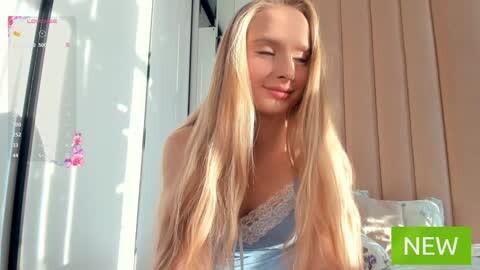 Media: Video of a young blonde woman with long hair, wearing a light blue lace bra, standing indoors in a brightly lit room with vertical blinds.