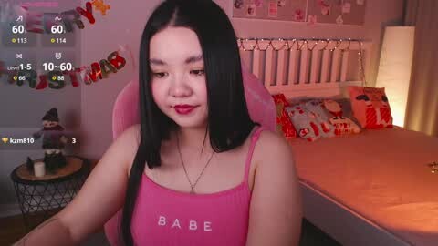 Media: Video of an Asian woman with straight black hair, wearing a pink \"Babe\" tank top, seated in a pink gaming chair in a cozy, warmly lit bedroom with Christmas decorations and \"Merry Christmas\" banner.