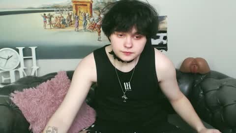 Media: Video of a pale-skinned, black-haired young man in a black sleeveless shirt, sitting on a gray couch with a pink pillow, wearing a black choker. Background features a poster and a large, realistic, brownish anatomical model of a female torso.