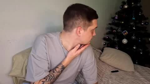 Media: Video of a young man with short dark hair, wearing a gray T-shirt and a silver necklace, lying on a bed with a beige pillow and a striped blanket, gazing at a decorated Christmas tree in the background.