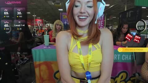 Media: Video of a young woman with red hair, wearing a yellow crop top, smiling at a virtual reality game booth in a crowded, vibrant indoor venue.