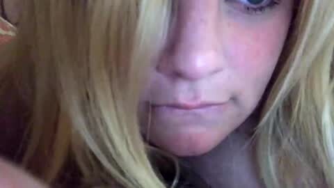 Video of a close-up of a young girl with fair skin and blonde hair, partially obscuring her face. She has a subtle blush on her cheeks and a slight smile. The background is blurred and indistinct.