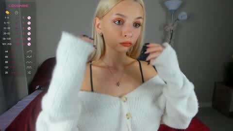 Media: A video of a young, fair-skinned woman with blonde hair, wearing a white off-shoulder cardigan and black bra, adjusting it in a bedroom with a red bedspread, a lamp, and a pink nightstand.