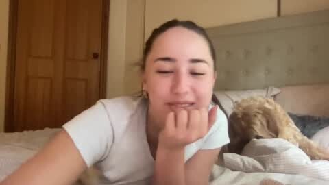 Media: Video of a woman with brown hair, fair skin, and closed eyes, lying on a bed, fingers to mouth. Background includes a beige headboard, wooden door, and rumpled bedding.