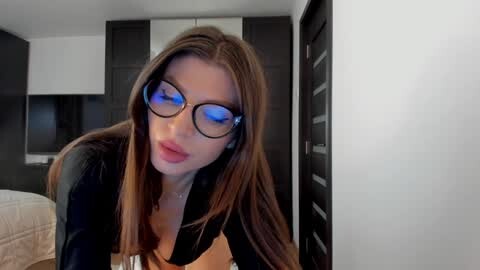Media: Video of a slender, fair-skinned woman with long brown hair, wearing black-rimmed glasses and a black top, leaning forward in a modern bedroom with dark wooden furniture and a TV.