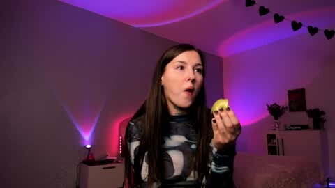 Media: Video of a young woman with long brown hair, wearing a patterned shirt, holding a lemon, in a dimly lit room with purple and pink LED lights.