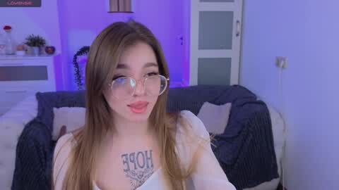 Media: Video of a young woman with long brown hair, fair skin, and large glasses, wearing a white top with \"HOPE\" tattooed on her chest, sitting on a sofa in a softly lit living room.
