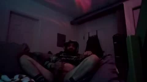 Media: A dimly-lit, grainy video of a man with dark hair, wearing a black hoodie, reclining on a bed with his legs spread, in a dimly lit, cluttered room with red and purple lighting.