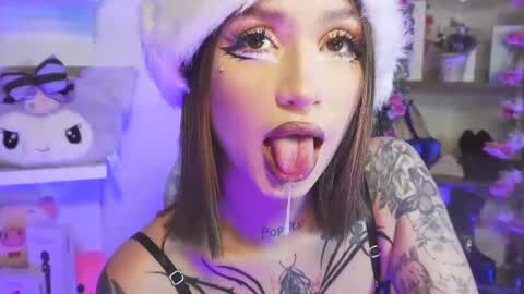 Media: Video of a young woman with light skin, dark hair, and tattoos, wearing a Santa hat and black lingerie, sticking out her tongue with drool. Background includes a stuffed animal and colorful, cluttered room.