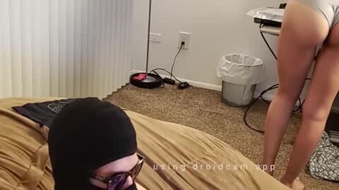 Media: Video: A man in a black beanie, glasses, and beige shirt, bending over a woman in white lingerie, exposing her buttocks, in a cluttered, carpeted bedroom with blinds and a laundry basket.