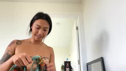 Media: Video of a topless woman with medium brown skin and black hair, holding a colorful piece of cloth, tattoos on her arms, in a bright, minimalist room.