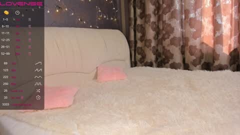 Media: Video of a cozy bedroom with a cream-colored tufted headboard, pink pillows, and a fluffy white bedspread. Background features patterned curtains and a nightstand. The image has a \"LoveSense\" logo overlay.