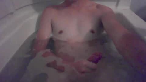 Media: Video of a fair-skinned, nude male with small breasts, sitting in a white bathtub filled with soapy water. He holds a pink object in his right hand.