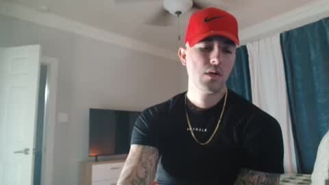 Media: A video of a young, pale-skinned man with a red baseball cap, black t-shirt, and multiple tattoos, standing in a modern bedroom with white walls, dark curtains, and a flat-screen TV.