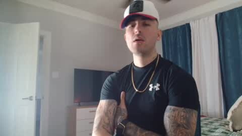 Media: Video of a muscular, tattooed man with a white baseball cap, red visor, and black Under Armour shirt, giving a thumbs-up in a simple bedroom with a flat-screen TV, white curtains, and navy blue drapes.