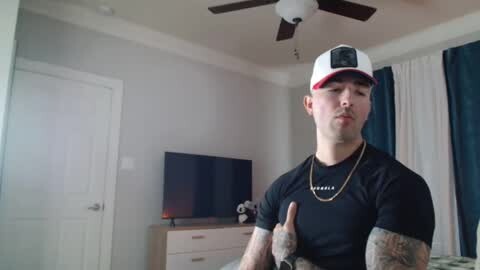 Media: Video of a young man with tattoos, wearing a black shirt, white cap, and necklace, giving a thumbs-up in a modern bedroom with a TV, dresser, and blue curtains.