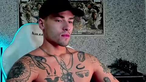 Media: Video of a shirtless, muscular man with light skin and tattoos on his chest, wearing a black cap, sitting in a white gaming chair, surrounded by a collage of dollar bills and a chessboard.