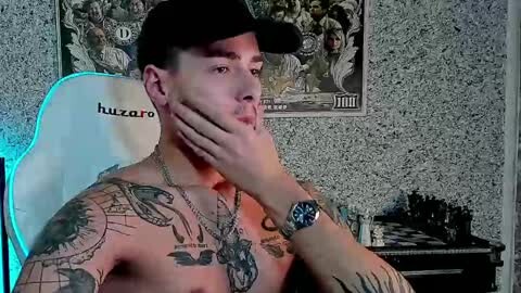 Media: Video of a shirtless, tattooed man with short black hair, wearing a black cap and a chain necklace, seated in a white \"huzro\" gaming chair, with a chessboard and a poster in the background.