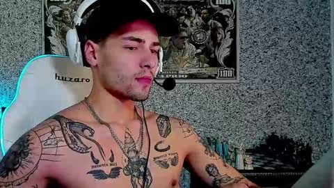 Media: Video of a shirtless, muscular man with light skin and multiple tattoos, wearing a black cap and headphones, sitting at a white gaming chair, in a dimly-lit room with a checkered chessboard and a poster on the wall.